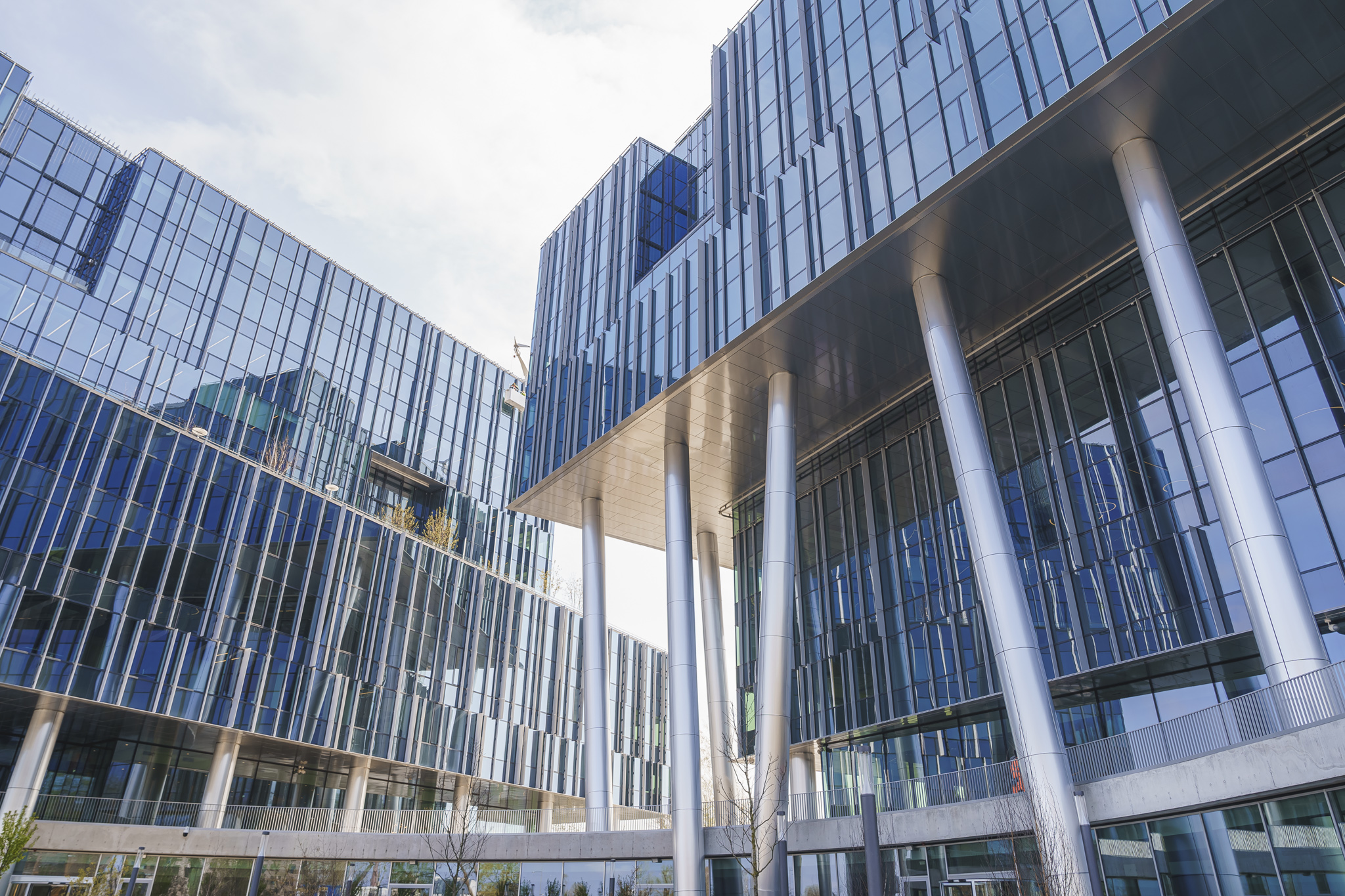 Modern blue-glass headquarters with silver pillars above a paved plaza, white outdoor seating, and young trees.