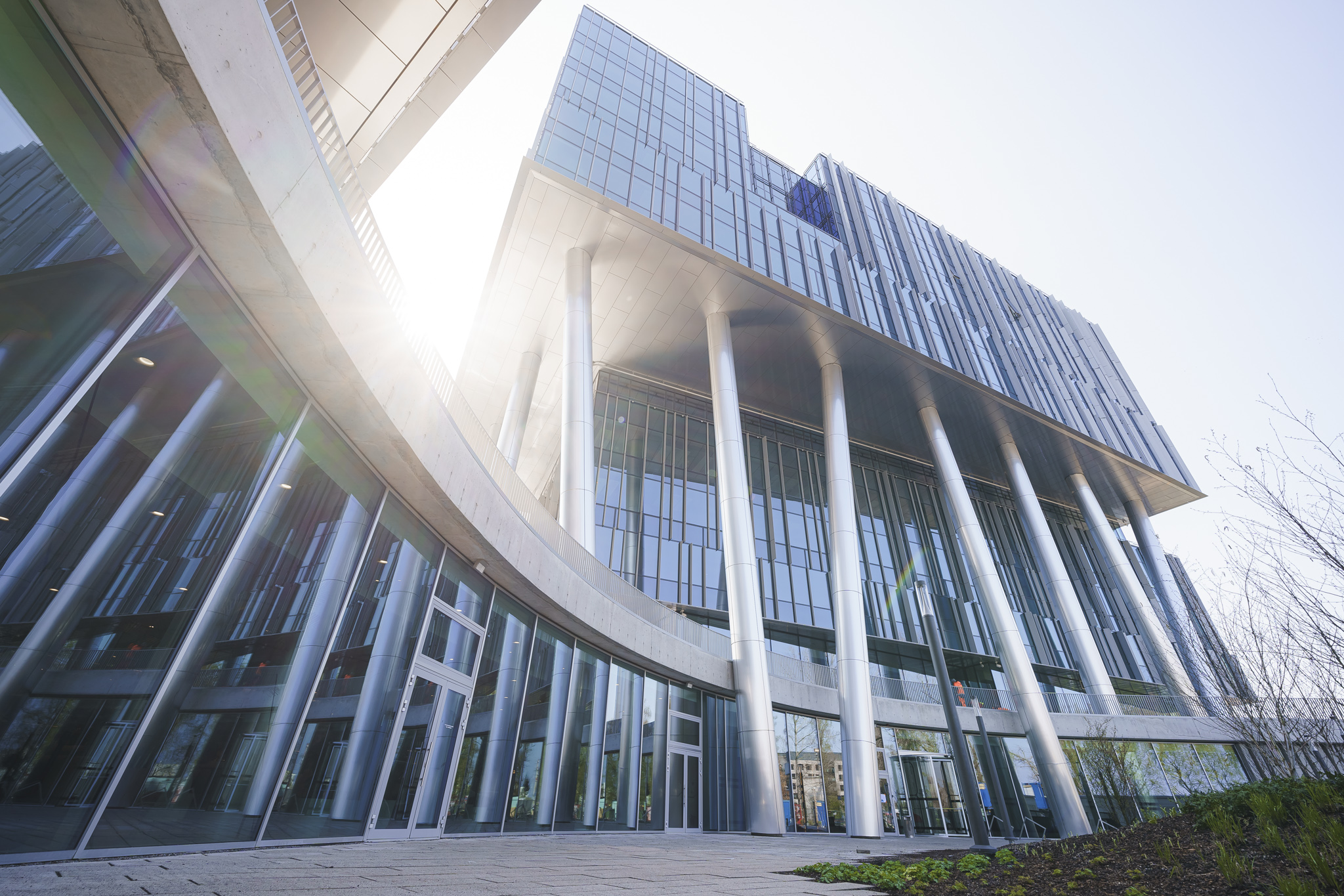Low-angle view of a curved glass office facade and suspended upper floors with bright sun flare against a clear sky.