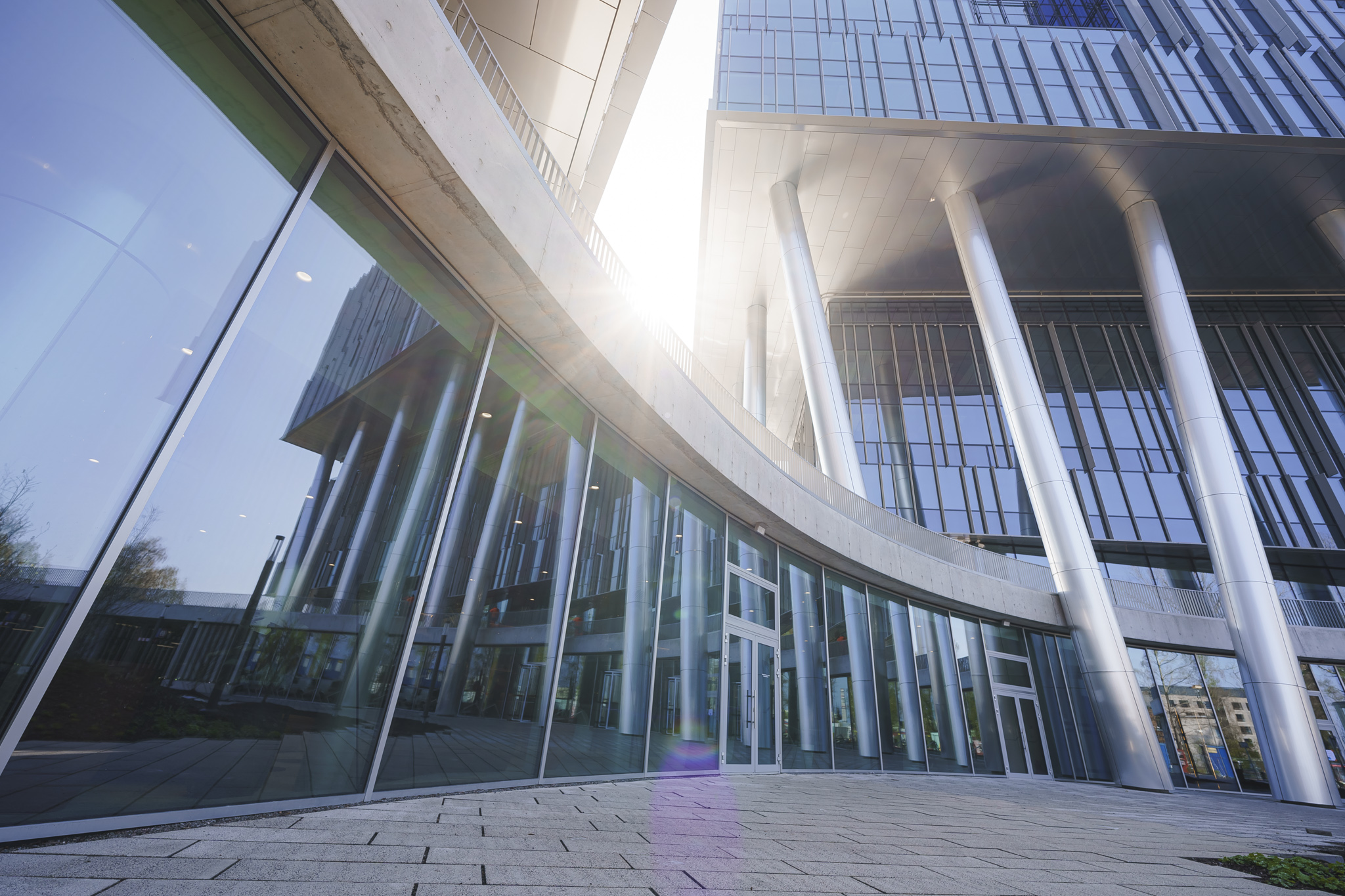 Low-angle view of two contemporary glass towers with silver structural pillars under a bright sky.